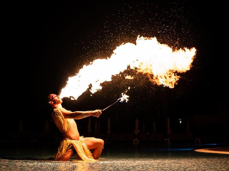 Mann entzündet Feuerstoß aus dem Mund, sitzt am Rand eines Wassers mit dunklem Hintergrund.