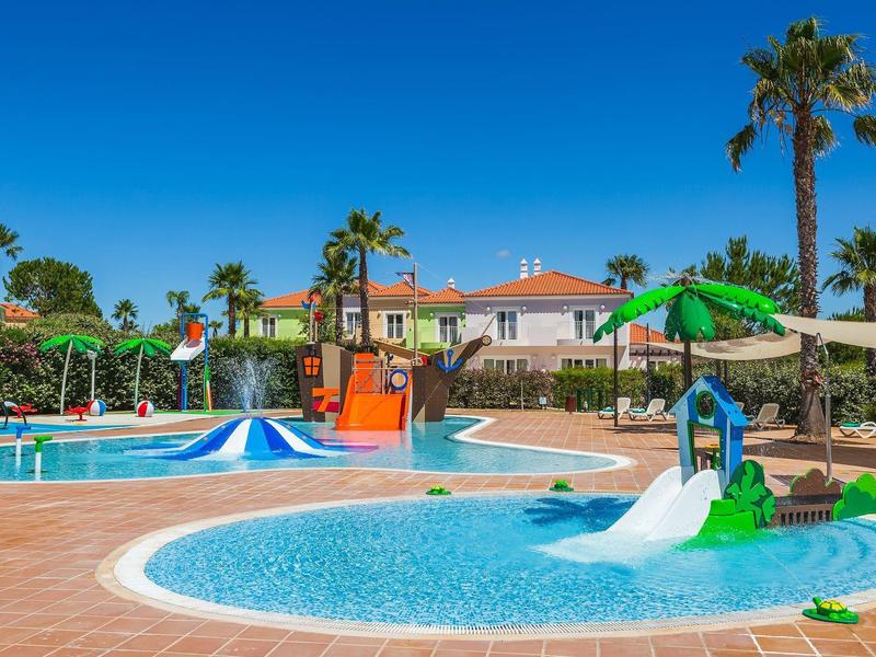 Großer Poolbereich mit Wasserrutschen, Palmen und blauen Sonnenschirmen unter klarem Himmel.