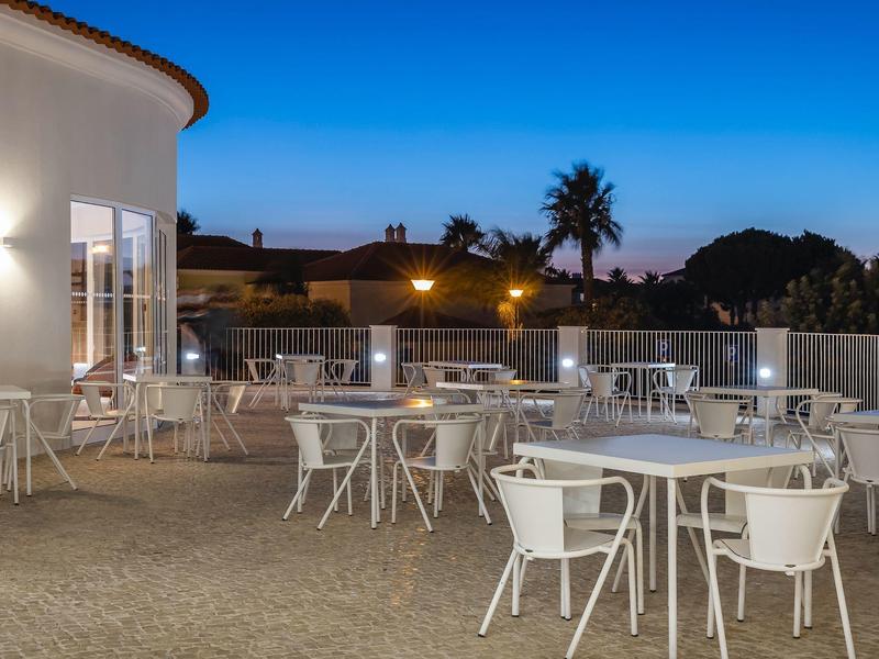 Leere Terrasse mit weißen Tischen und Stühlen bei klarem Abendhimmel, Palmen im Hintergrund.