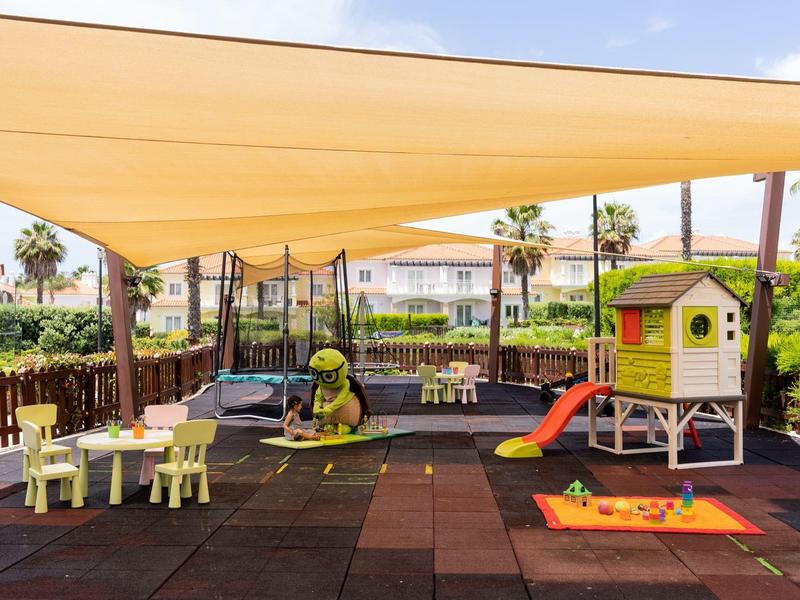 Bunter Kinderspielplatz mit Rutsche, Sandkasten, Tisch und Stühlen unter sonnigem Himmel.