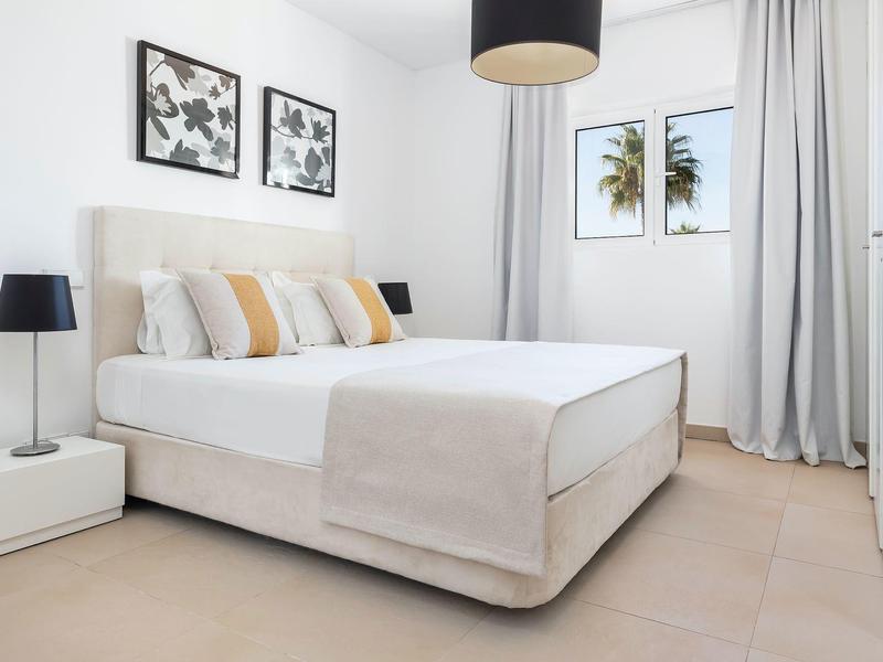 Helles Schlafzimmer mit großem Bett, beigen Vorhängen, Nachttischen und zwei Wandbildern.