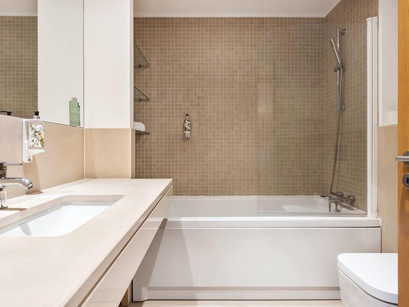 Modernes Badezimmer mit rechteckiger Badewanne, beige Fliesen und Waschbecken auf langer Ablagefläche.