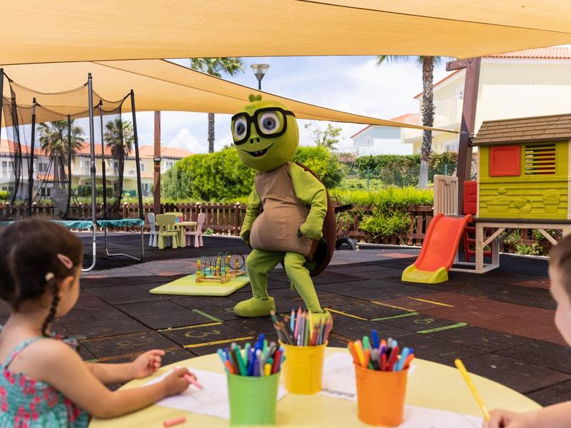 Kinder sitzen an einem Tisch mit Farben, vor ihnen steht eine Person im grünen Dinosaurierkostüm im Freien.
