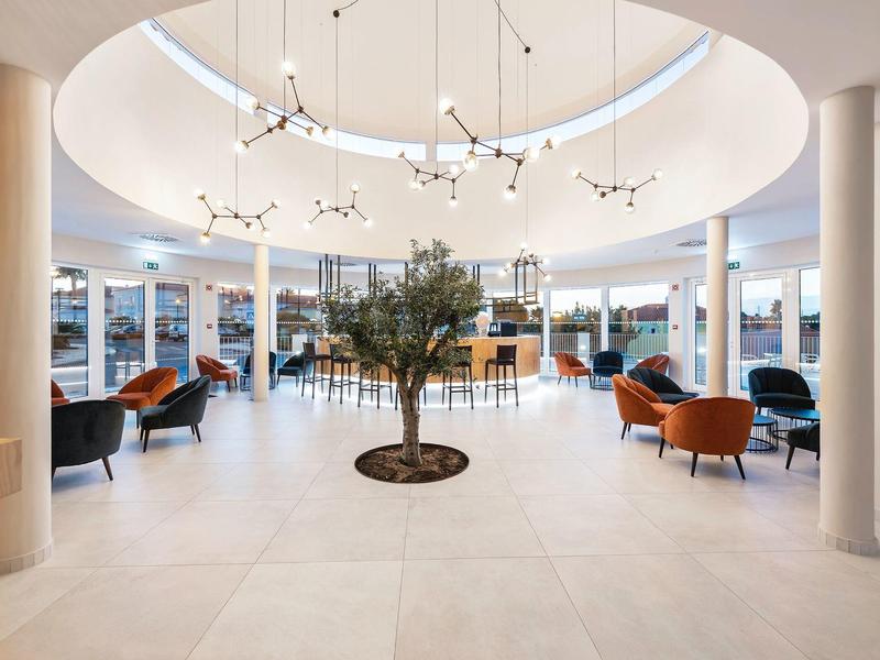 Großer heller Raum mit runden Säulen, zentralem Baum und modernen Stühlen in warmen Farben.