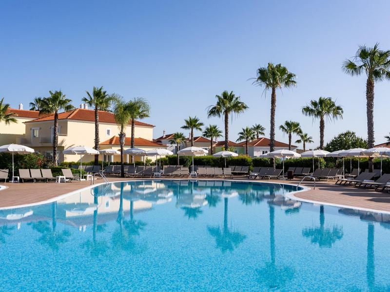 Großer Swimmingpool mit Palmen, Liegestühlen und Sonnenschirmen in einer sonnigen Hotelanlage.