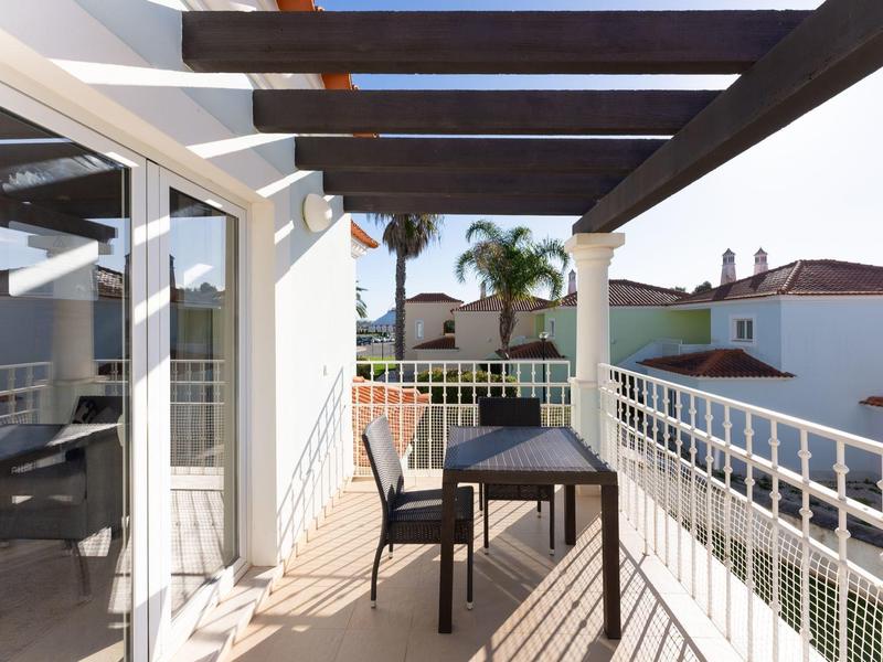 Balkon mit Tisch und zwei Stühlen, überdacht mit dunklen Holzbalken, in sonniger Umgebung.