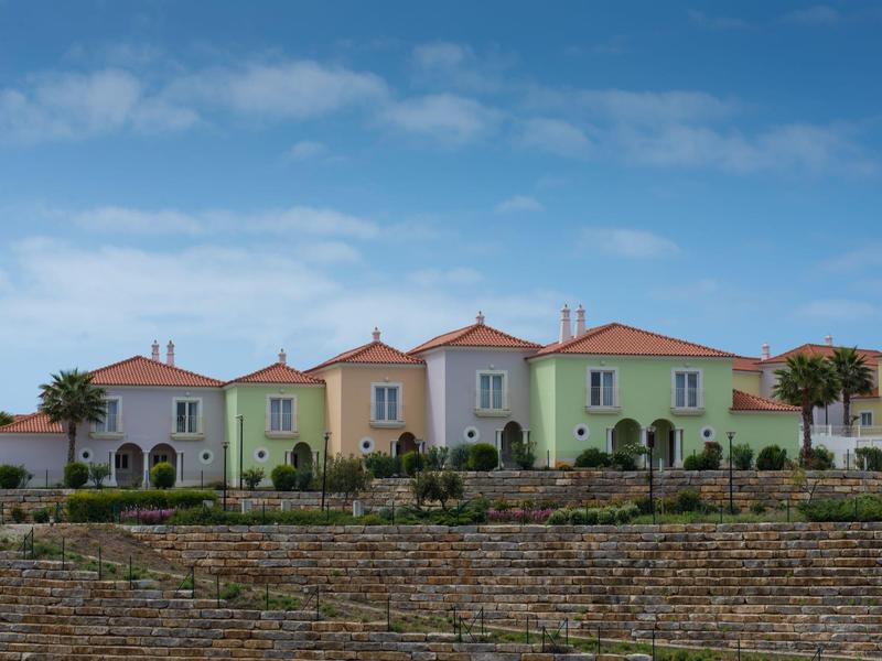 Reihe farbiger, einstöckiger Häuser mit roten Dächern vor blauem Himmel und Steinmauer im Vordergrund.