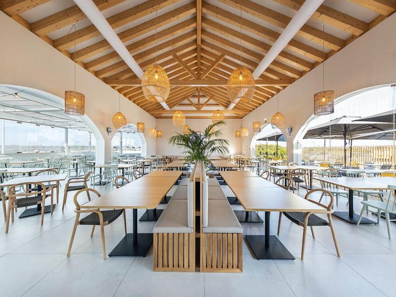 Modernes, helles Restaurant mit Holzdecke, großen Fenstern, minimalistischen Tischen und Stühlen.