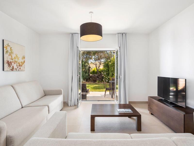 Helles Wohnzimmer mit weißen Wänden, beigen Sofas, braunem Couchtisch und Fernseher vor Gartentür.