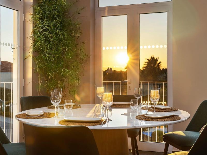 Runder Esstisch mit Gläsern und Tellern vor großen Fenstern, Sonnenuntergang im Hintergrund.