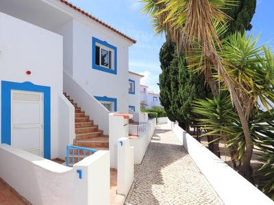Weiße Häuser mit blauen Fensterrahmen und Türen, Treppe, heller Kiesweg und Palmen im Sonnenlicht.