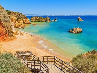 Ein heller Sandstrand mit Felsen im türkisblauen Meer und Holzstegen im Vordergrund unter blauem Himmel.