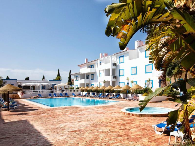 Heller Außenpool mit blauen Liegestühlen neben weißem Hotelgebäude und Palmen bei klarem Himmel.