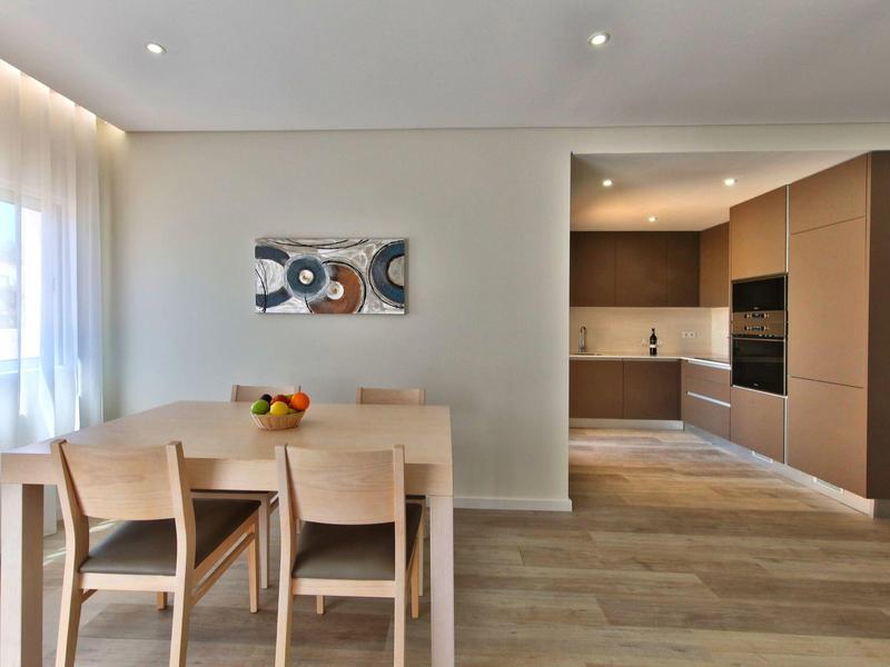 Moderner Essbereich mit hellem Holztisch, vier Stühlen, Küchenzeile und großem Fenster mit Gardine.