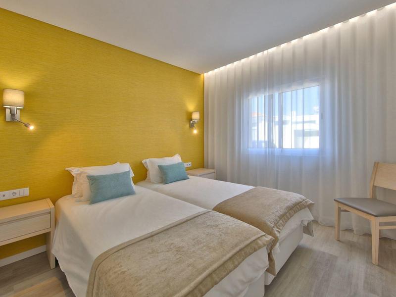 Zweibettzimmer mit gelber Wand, zwei Betten, weißen Vorhängen und schlichtem Holzstuhl.