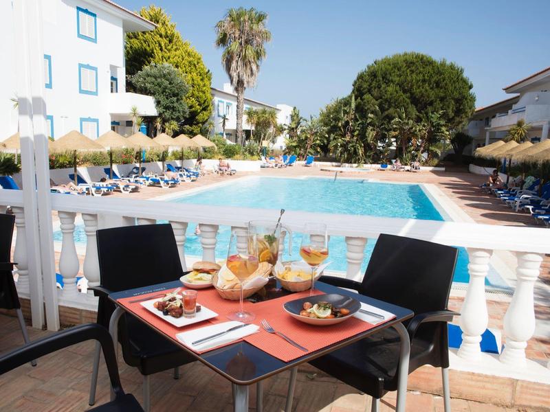 Sonniger Poolbereich mit blauem Wasser, Liegestühlen und gedecktem Tisch mit Speisen und Getränken.