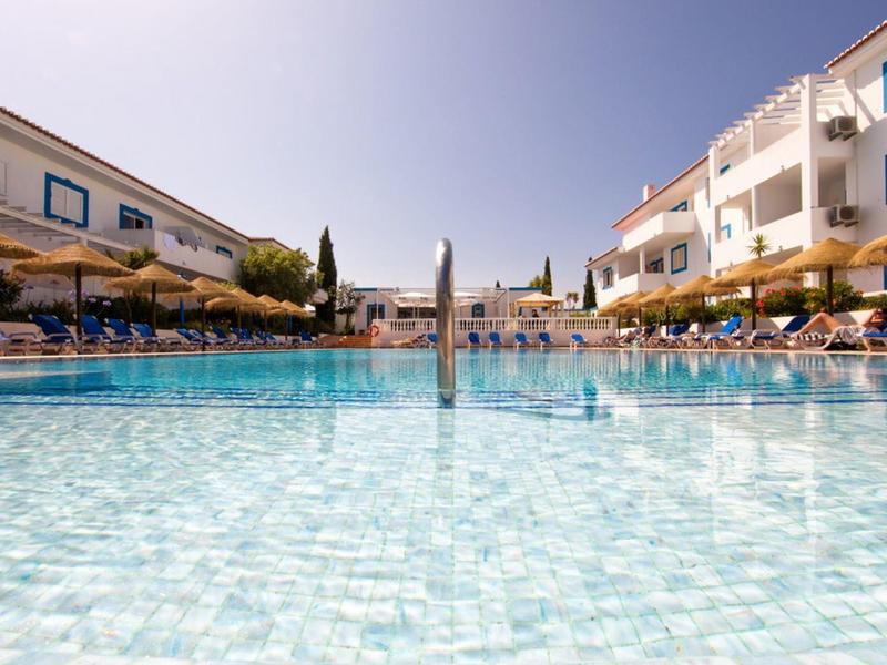 Großer klarer Pool mit Liegestühlen und Sonnenschirmen zwischen zwei weißen Gebäuden bei blauem Himmel.
