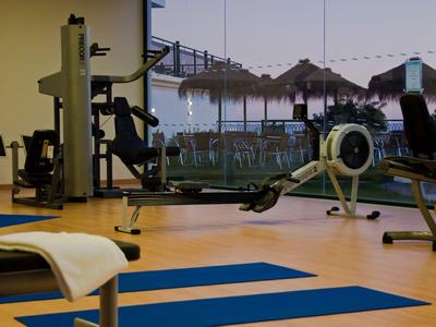 Innenansicht eines Fitnessraums mit Geräten und Blick auf eine Terrasse mit Sonnenschirmen.