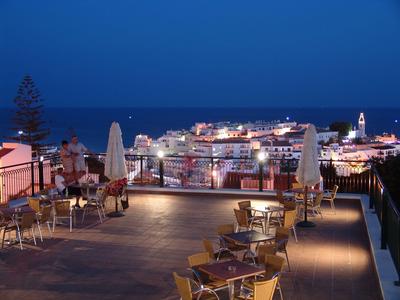 Abendlicher Blick auf eine Terrasse mit Tischen und Stühlen, im Hintergrund Hafenlichter und Meer.