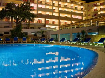 Beleuchteter Hotelpool bei Nacht mit Liegestühlen und Balkonen im Hintergrund.