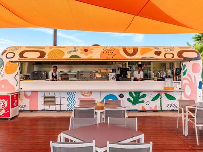 Bunte Bar mit tropischen Motiven und Sitzgelegenheiten unter einem orangen Sonnenschirm.