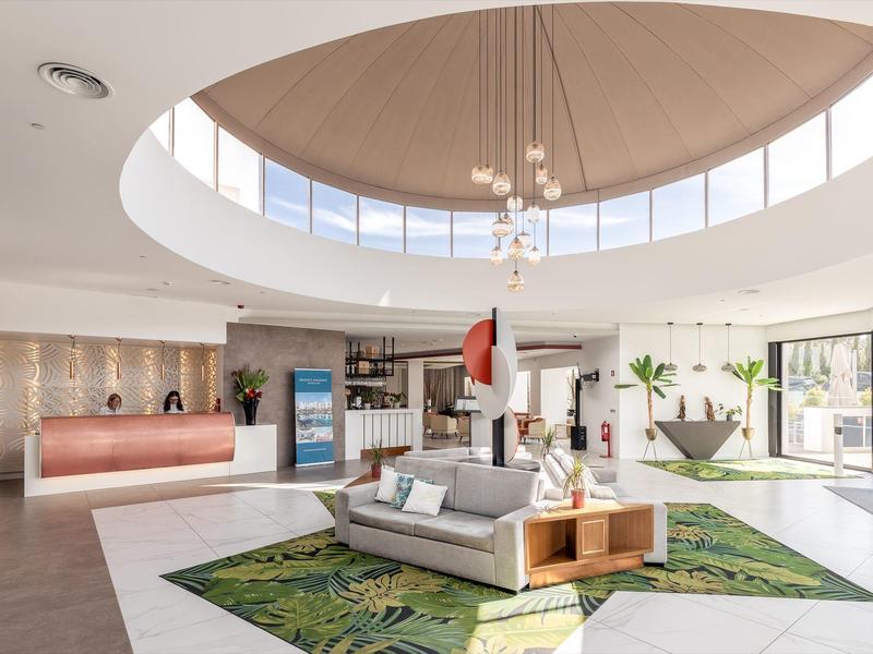 Helle, runde Hotellobby mit großem Fensterkreis, grünem Teppich und moderner Sitzgruppe in der Mitte.