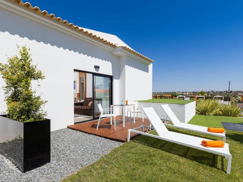 Modernes weißes Haus mit Holzterrasse, zwei weißen Sonnenliegen und grünem Rasen im Garten.