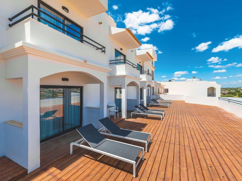 Eine Terrasse mit Holzboden und schwarzen Liegestühlen vor weißen Balkonen unter blauem Himmel.