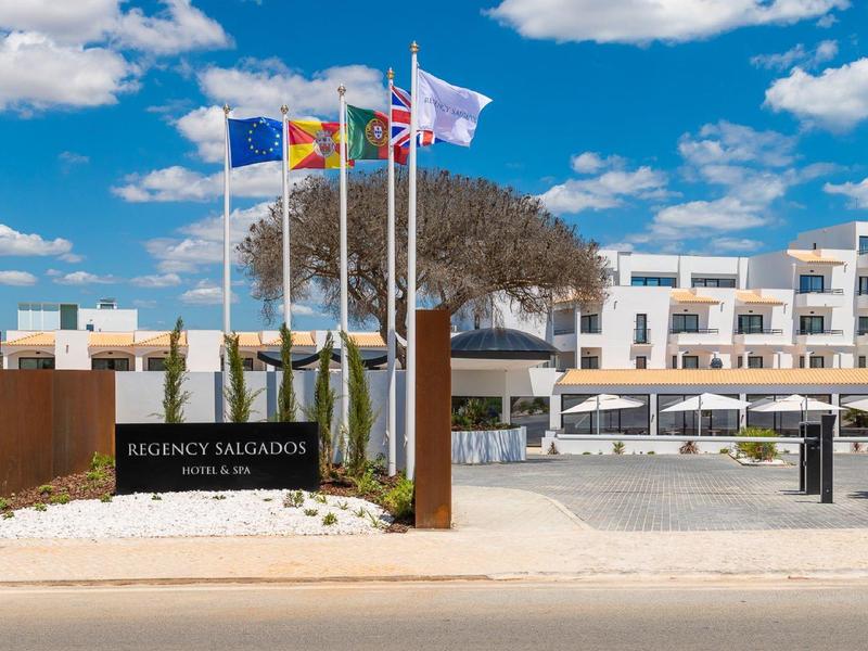 Hotelanlage mit weißen Gebäuden, Fahnenmasten, klar blauem Himmel und Sandfläche im Vordergrund.