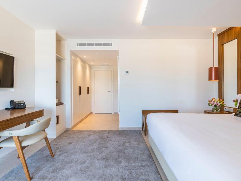 Helles Hotelzimmer mit großem Bett, Schreibtisch, Stuhl und Flachbildfernseher an weißer Wand.