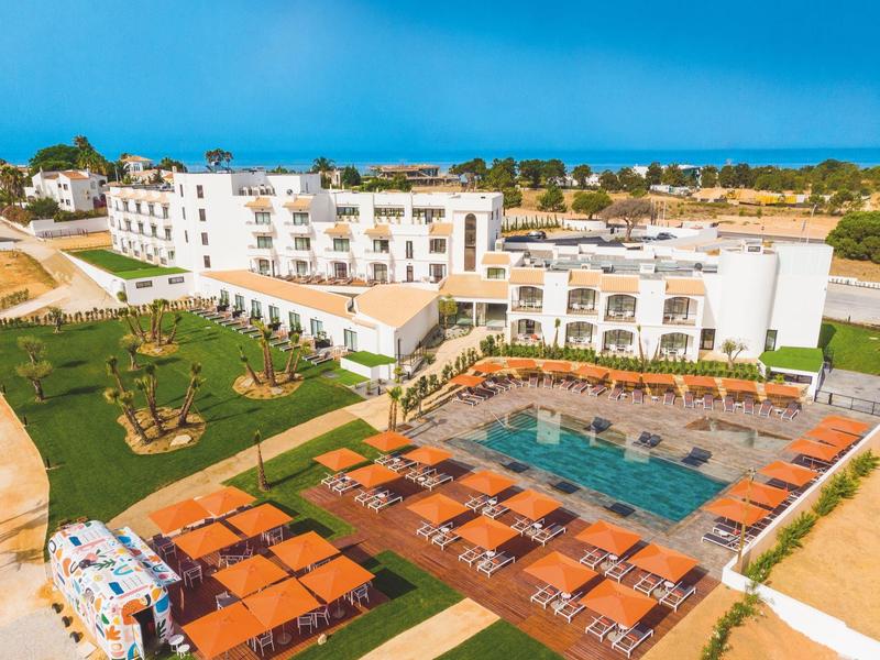 Großes weißes Hotel mit Pool, Sonnenschirmen, grüner Wiese und blauem Himmel im Hintergrund.