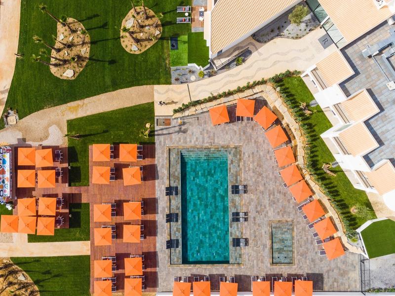 Vogelperspektive eines Pools mit orangen Liegestühlen und grünen Rasenflächen, umgeben von Gebäuden.