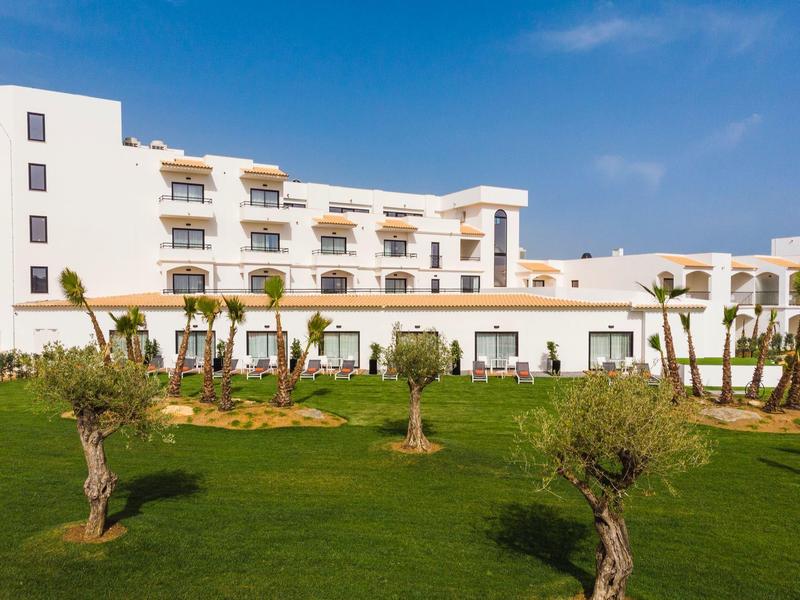 Weißes Hotelgebäude mit Balkonen, grünem Rasen und kleinen Bäumen bei blauem Himmel.