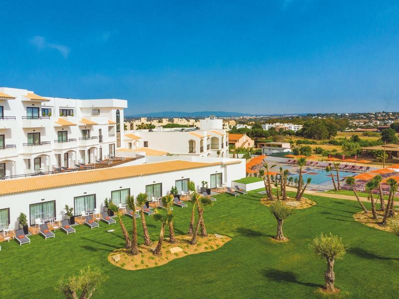 Hotelgebäude mit mehreren Stockwerken, grüner Rasen, Palmen und Pool bei klarem Himmel.