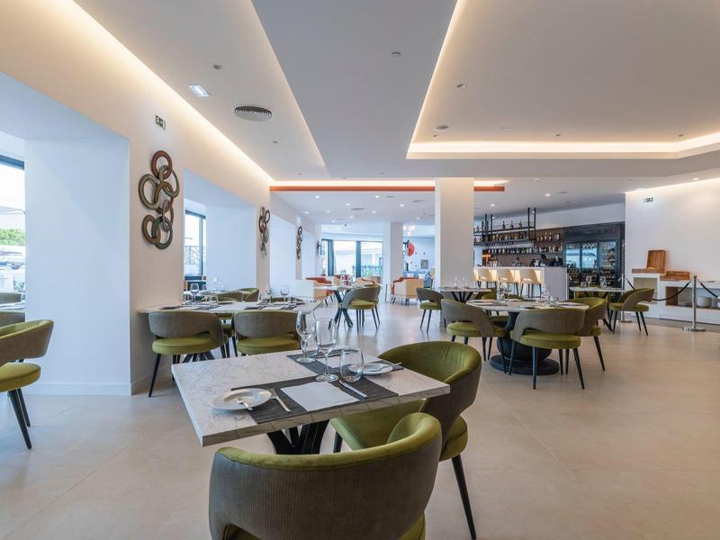 Moderner, heller Gastraum mit grünen Stühlen, weißen Tischen und großem Fensterfront.