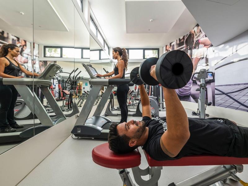 Mann trainiert mit Hantel auf Bank, Frau läuft auf Laufband, spiegelnde Wand im Fitnessraum.