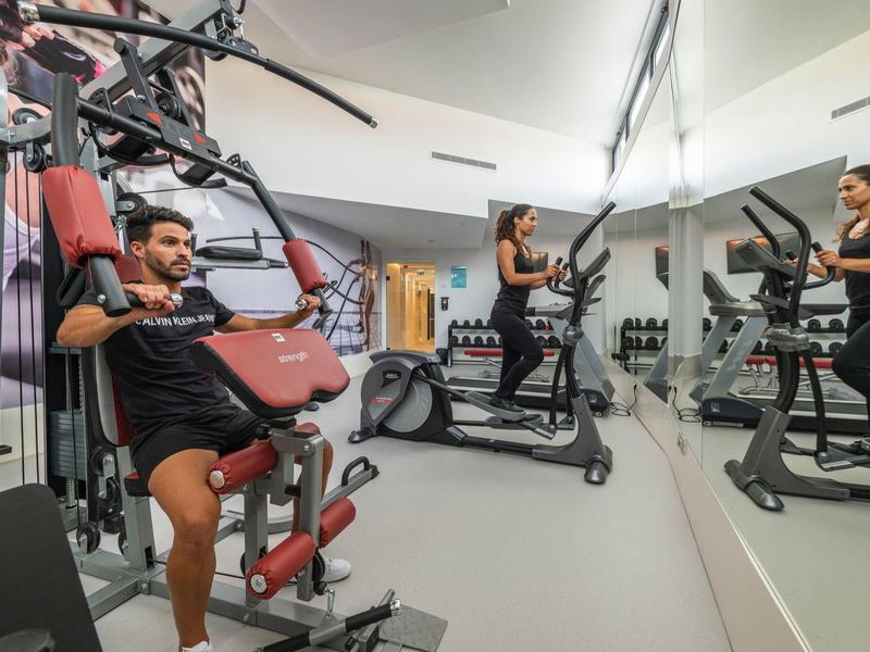 Fitnessraum mit zwei Personen, eine trainiert an Kraftgerät, die andere auf Laufband vor Spiegel.