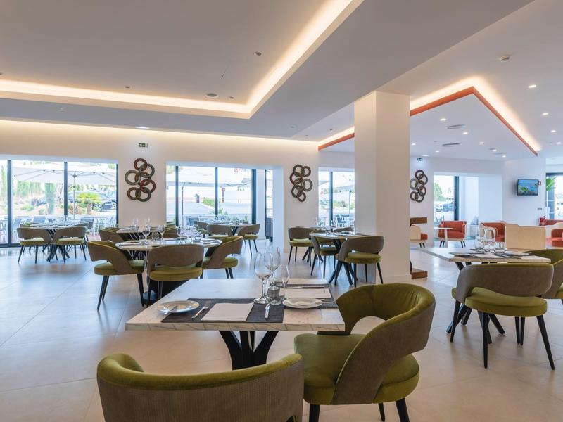 Moderner, heller Speisesaal mit großen Fenstern, grünen Stühlen und runden Tischen in hellem Ambiente.