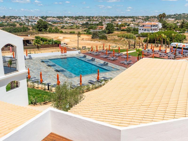 Blick auf ein großes Schwimmbecken mit roten Sonnenschirmen und Liegen, umgeben von weißen Gebäuden.