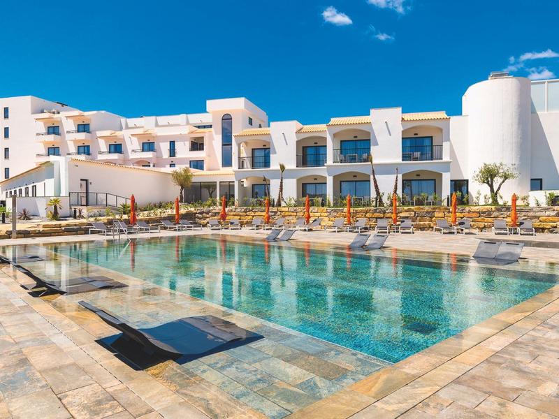 Moderner Hotelkomplex mit großem rechteckigem Pool und vielen Sonnenliegen am Beckenrand.