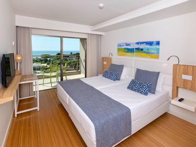 Helles Hotelzimmer mit Doppelbett, Balkon und Blick auf das Meer bei sonnigem Wetter.