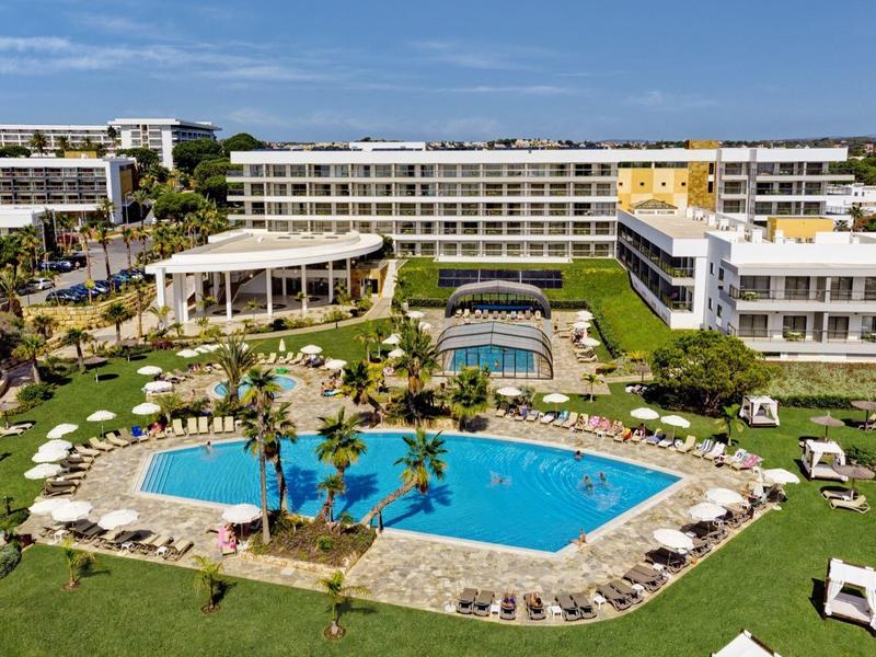 Hotel mit großem Pool, Pavillon und Liegestühlen auf einer grünen Rasenfläche bei klarem Himmel.