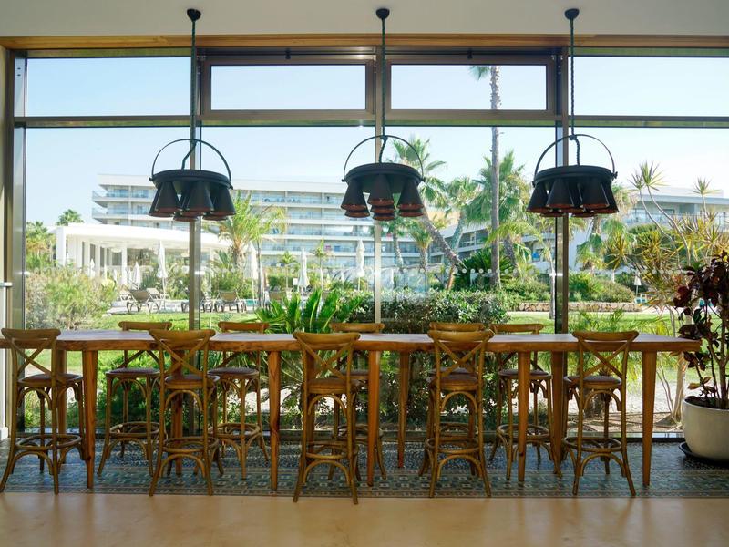 Moderner Essbereich mit Holzstühlen und Tischen vor großen Fenstern mit Blick auf tropische Pflanzen.