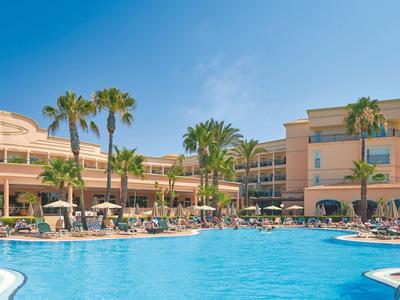 Großer Außenpool mit Liegestühlen und Palmen vor einem Hotelgebäude am sonnigen Tag.