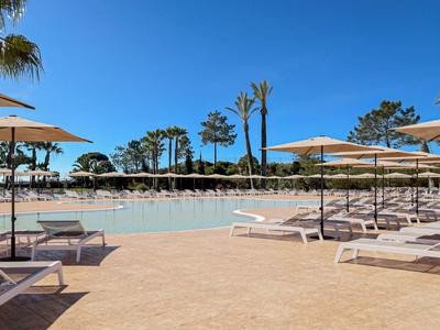 Sonniger Hotelpool mit Liegestühlen und Palmen unter klarem blauem Himmel.