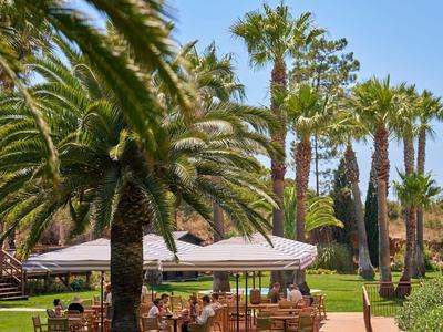 Grüner Garten mit Palmen, Tischen und Sonnenschirmen unter klarem blauem Himmel.