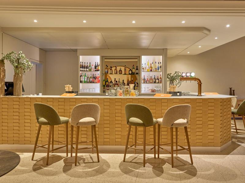 Moderner Hotelbarbereich mit Holzbar und vier hohen Stühlen in gedeckten Farben.