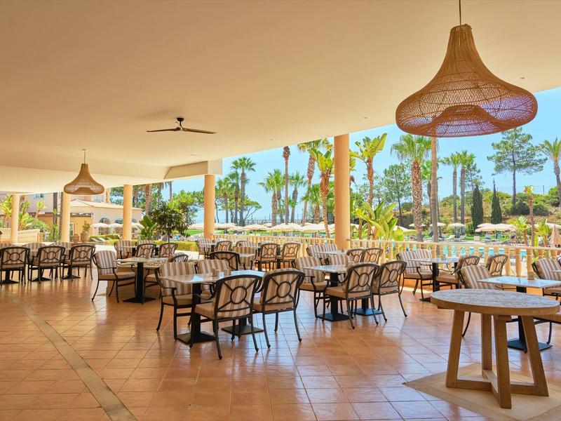 Offener Hotelbereich mit Tischen und Stühlen, Palmen und Pool im Hintergrund.