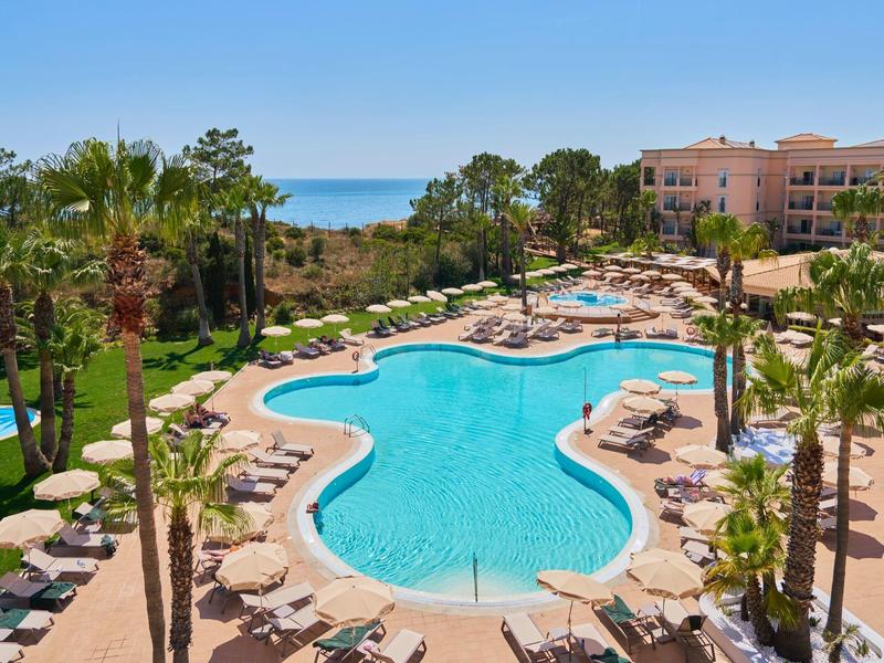 Großer Außenpool mit Sonnenschirmen und Palmen neben einem Hotel am Meer.