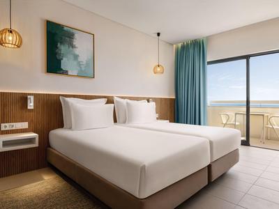 Modernes Hotelzimmer mit Doppelbett, Holzvertäfelung, türkisfarbenen Vorhängen und Balkon mit Meerblick.